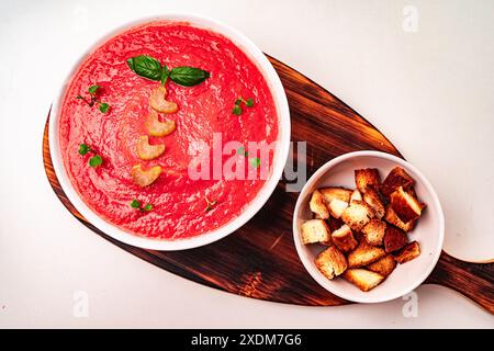 Deliziosa zuppa di crema di pomodoro con sedano e basilico in una ciotola bianca su un asse di legno con crostini su sfondo chiaro. Foto di alta qualità Foto Stock