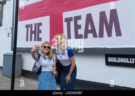 Londra, Regno Unito, 23 giugno 2024. Due tifosi scozzesi si mettono in posa per un selfie di fronte a un cartellone God Save the Team St George's a Finsbury Park. Il design del cartellone dell'artista Corbin Shaw in una varietà di luoghi diversi cerca di mettere in discussione ciò che l'Inghilterra rappresenta oggi. Credito: Fotografia dell'undicesima ora/Alamy Live News Foto Stock