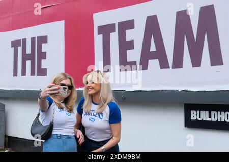 Londra, Regno Unito, 23 giugno 2024. Due tifosi scozzesi si mettono in posa per un selfie di fronte a un cartellone God Save the Team St George's a Finsbury Park. Il design del cartellone dell'artista Corbin Shaw in una varietà di luoghi diversi cerca di mettere in discussione ciò che l'Inghilterra rappresenta oggi. Credito: Fotografia dell'undicesima ora/Alamy Live News Foto Stock