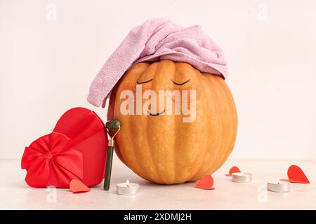 Zucca con viso disegnato, toppe sotto gli occhi, rullo di giada e regalo a forma di cuore su sfondo chiaro Foto Stock