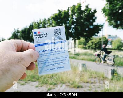 Strasburgo, Frnace - 7 giugno 2024: Mano che tiene in mano una carta elettorale francese carte Electorale all'aperto, con un ciclista sullo sfondo e alberi che fiancheggiano A. Foto Stock