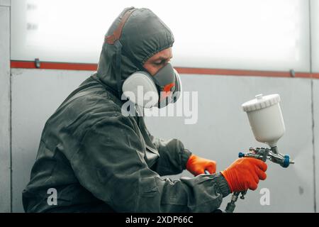 Pittore maschio in maschera respiratoria e tuta protettiva verniciatura auto con pistola a spruzzo in servizio auto Foto Stock