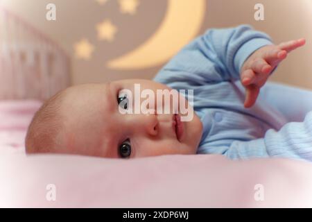 Il tranquillo bambino si stende su un letto accanto a una culla, circondato da un'atmosfera accogliente e dai toni delicati. Foto Stock