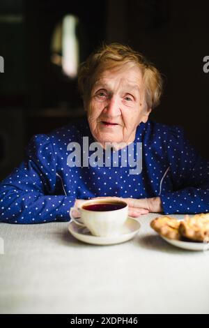 Una donna anziana si siede a un tavolo, bevendo una tazza di tè. Il ritratto cattura il suo sorriso dolce e il calore di un momento tranquillo a casa. Foto Stock