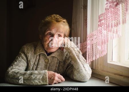 Una donna anziana in un maglione si siede accanto a una finestra, guardando l'esterno con un'espressione serena, catturando un momento di riflessione pacifica. Foto Stock