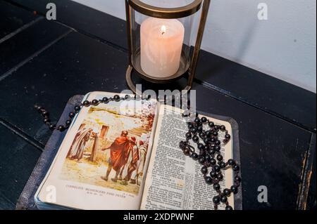 Una Bibbia, aperta sulla storia di Cornelio, adagiata su una credenza di legno con perline di rosario nere sopra di essa e una candela dietro di essa Foto Stock