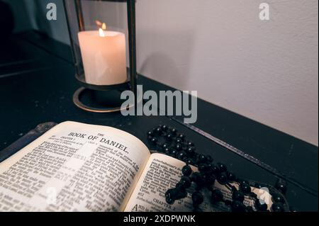 Una Bibbia, aperta al Libro di Daniele, adagiata su una credenza di legno con perline nere di rosario sopra di essa e una candela dietro Foto Stock