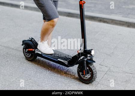 Immagine corta dell'uomo in pantaloncini corti e sneakers che saltano su uno scooter elettrico mentre si trova in città. Ecologia e modalità di trasporto ecologiche pulite Foto Stock