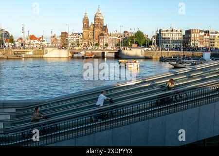 I ciclisti spingono le loro biciclette verso l'alto dal parcheggio sotterraneo per biciclette della stazione Centraal, con il canale e la basilica di San Nicola sullo sfondo, ad Amsterdam, Paesi Bassi. Foto Stock