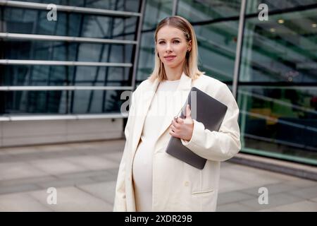 Una bella donna d'affari bionda incinta con un abito da lavoro bianco e un blazer si trova all'aperto con un laptop vicino a un edificio aziendale, uno show-cas Foto Stock