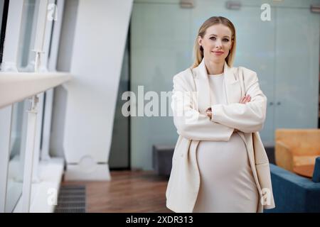 Bella donna d'affari bionda incinta con un abbigliamento elegante e alla moda un abito bianco e un blazer che guarda la fotocamera in un'azienda Foto Stock