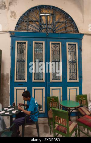 Tunisi, Tunisia. 7 maggio 2024. Un giovane tunisino gioca a carte e beve il tè accanto a una tradizionale porta ad arco blu in un caffè nella Medina di Tunisi, in Tunisia. (Credit Image: © John Wreford/SOPA Images via ZUMA Press Wire) SOLO PER USO EDITORIALE! Non per USO commerciale! Foto Stock