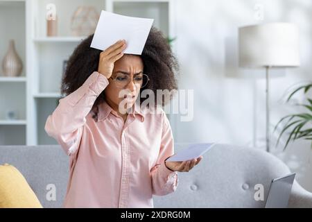 Donna frustrata in abiti casual, in possesso di documenti finanziari, che soffre di stress nella gestione delle bollette a casa. Emotivamente sopraffatti dall'ambiente moderno del soggiorno. Concetto di finanza personale, stress Foto Stock