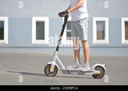 Un uomo con abbigliamento sportivo casual guida uno scooter elettrico sullo sfondo di un edificio. Vista laterale. Il concetto di ecologia e stile di vita sano in città Foto Stock
