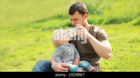 Il piccolo papà dà da mangiare al bambino seduto sul biberon in erba nel soleggiato parco estivo Foto Stock