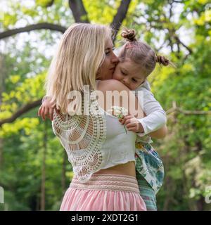 Mamma e figlia nel parco. Mamma con capelli biondi in una maglietta bianca a rete e una gonna rosa tiene una bambina delicatamente tra le braccia. La figlia l'ha fatto Foto Stock