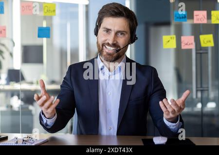 Un uomo d'affari sorridente che indossa tuta e cuffia per riunioni virtuali in un ufficio moderno. Comunicazione professionale sicura in remoto grazie alla tecnologia di videoconferenza e al lavoro interattivo Foto Stock