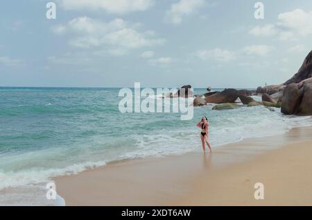 Una donna solitaria in un bikini nero si trova su una spiaggia sabbiosa, di fronte all'oceano. Le onde ondulate si infrangono sulla riva e sono visibili grandi affioramenti rocciosi Foto Stock