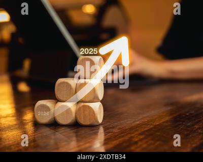 Aumenta la freccia da 2024 a 2025 anni su una pila di blocchi di legno a cubetti, scala il grafico di crescita aziendale su un tavolo di legno mentre la persona lavora con il computer. B Foto Stock