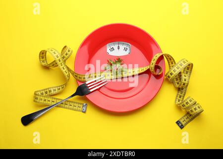 Piastra rossa con display analogico della bilancia, rucola, forchetta e metro su sfondo giallo, vista dall'alto Foto Stock