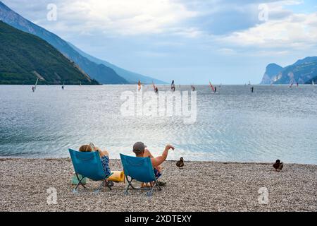 Torbole, Lago di Garda, Italia - 24 giugno 2024: due turisti in coppia su una sdraio sulla spiaggia del Lago di Garda in Italia guardando il panorama con windsurf e montagne *** Zwei Touristen als Pärchen in einem Liegestuhl am Strand vom Gardasee in Italien blicken auf das Panorma mit Windsurfern und Bergen Foto Stock