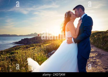 Ritratto dello sposo e della sposa che si baciano vicino all'oceano al tramonto dopo il matrimonio Foto Stock