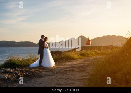 Marito e moglie contemplano un faro al tramonto vicino all'oceano dopo il matrimonio Foto Stock