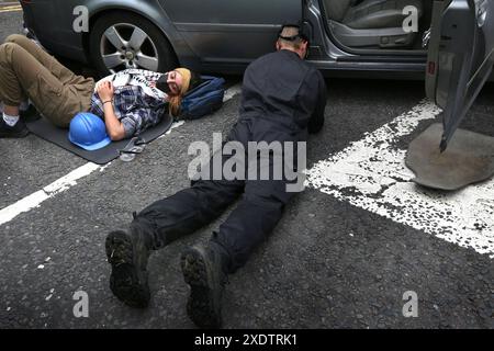 24 giugno 2024, Bristol, Inghilterra, Regno Unito: Un ufficiale di polizia specializzato guarda sotto la macchina per provare a vedere come gli attivisti sono connessi in modo che il suo team possa disaccoppiarli. I sostenitori di Palestine Action usano le chiuse per bloccare l'unico accesso veicolare alla compagnia di difesa israeliana, l'attività di Elbit Systems ad Aztec West, Bristol. Essi sostengono che le armi prodotte da Elbit nel Regno Unito sono usate dagli israeliani contro i palestinesi a Gaza e altrove. I bombardamenti israeliani a Gaza hanno ucciso oltre 35.000 palestinesi dall'ottobre 2023. Palestina Action è determinata a prendere di mira senza sosta Elbit e la loro pa Foto Stock