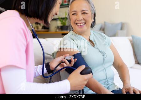 Infermiera femminile che controlla la pressione sanguigna di donna asiatica anziana, sorridendo sul divano Foto Stock