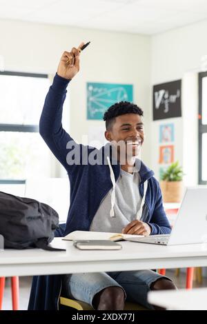 A scuola, uno studente maschio che alza la mano mentre usa il laptop in classe Foto Stock