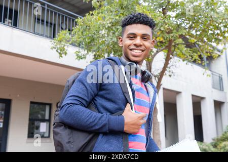 Studente maschio sorridente con zaino e cuffie in piedi fuori dall'edificio scolastico Foto Stock