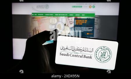 Persona che detiene un telefono cellulare con il logo dell'istituto finanziario Saudi Central Bank (SAMA) di fronte alla pagina web. Mettere a fuoco il display del telefono. Foto Stock