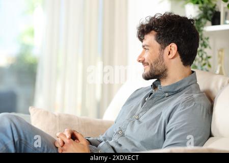 Uomo felice seduto su un divano che guarda da una finestra di casa Foto Stock