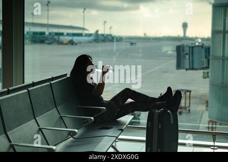 Donna che beve una bottiglia d'acqua mentre aspetta il volo seduto e rilassante su una panchina all'aeroporto. Foto Stock