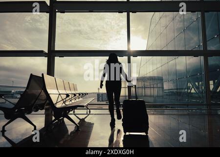 Una donna con la valigia cammina attraverso la sala delle partenze dell'aeroporto mentre i raggi del sole passano dalla finestra. Foto Stock