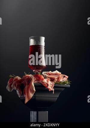 Bicchiere di vino rosso e prosciutto con rosmarino su un podio nero. Foto Stock