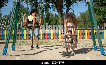 Madre e padre latini che fanno oscillare i loro figli in un parco giochi. Famiglia venezuelana con un bambino e un bambino Foto Stock
