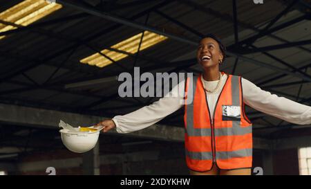 Ingegnere donna entusiasta che indossa il giubbotto di sicurezza che regge l'elmetto in cantiere Foto Stock
