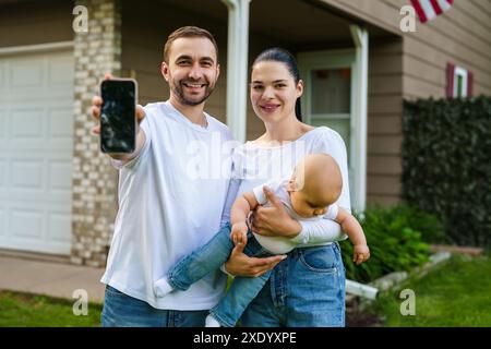 Giovane famiglia in piedi di fronte a casa all'aperto con lo schermo del telefono. Foto Stock