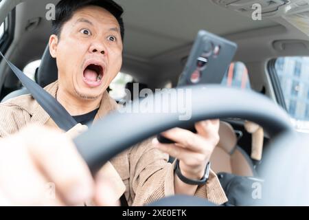 Un uomo sta guidando un'auto e guardando il suo cellulare. Sta urlando ed è in stato di shock. Foto Stock