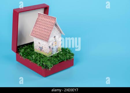 Una piccola casa è collocata all'interno di una scatola rossa. La casa è fatta di carta e si trova su un'area verde erbosa. La scatola è rossa e ha una finitura lucida. Il Foto Stock