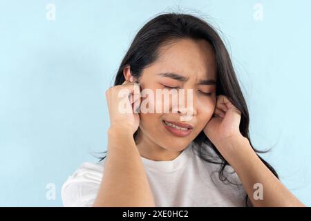 Giovane donna che soffre di dolore all'orecchio su sfondo azzurro. Stressata giovane ragazza etnica che tace l'orecchio, sentendo dolore disagio. Foto Stock