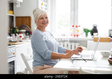 Donna matura che usa un notebook seduto al tavolo in cucina Foto Stock
