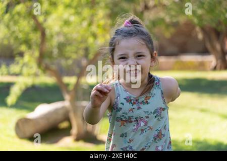 Una bambina con un volto malizioso corre verso la telecamera, facendo gesti divertenti in un parco soleggiato. Perfetto per catturare gioia, energia infantile e te Foto Stock