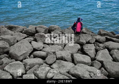 Persona seduta su rocce frangiflutti con protezione solare e attrezzatura da pesca, da sola. Foto Stock