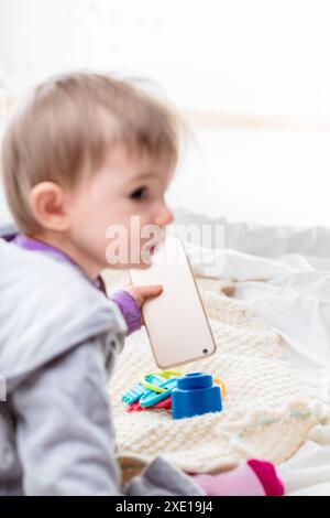 Bimba felice che gioca con uno smartphone su un comodo letto, perduta nella tecnologia, i suoi capelli biondi brillano su uno sfondo bianco. Meraviglia dell'infanzia in t Foto Stock