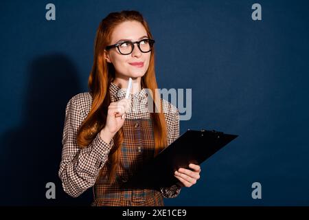 Foto di un vestito a quadri da donna dolce da sogno che scrive appunti con uno spazio in rilievo, sfondo di colore blu scuro isolato Foto Stock