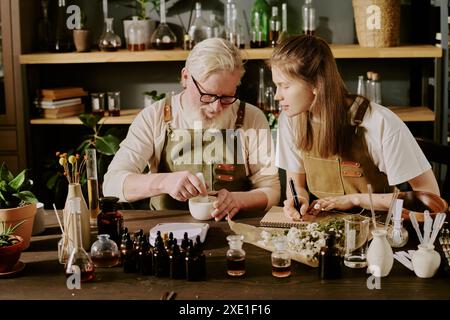 Profumiere maturo che utilizza mortaio e pestello in ceramica per schiacciare le erbe mentre il suo giovane stagista seduto accanto a lui e annotando questo processo Foto Stock