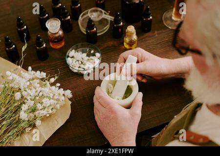 Un profumiere maturo che trita margherite essiccate in mortaio di ceramica con pestello di ceramica bianco, seduto alla scrivania con essenza diversa in bottiglie di vetro marrone Foto Stock