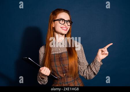 Foto di un vestito a quadri da donna brillante e sognante che regge gli appunti puntando verso uno spazio vuoto con sfondo blu scuro isolato Foto Stock